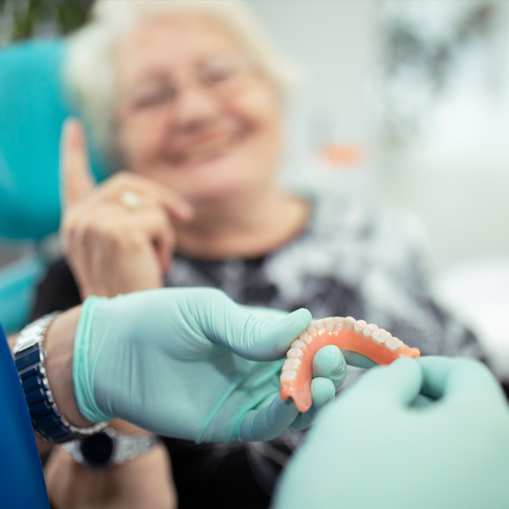 blurred image of an old lady in the background and doctor's hand holding lower set of dentures in her hand in foreground